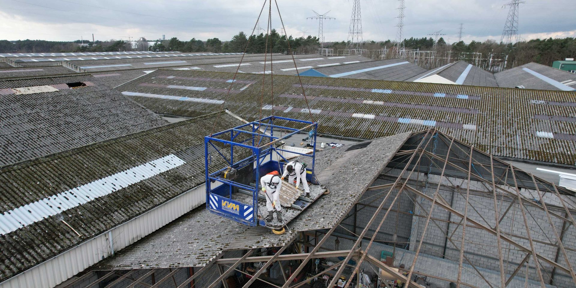 Twee arbeiders in witte beschermkleding op een hijskooi verwijderen dakplaten boven een industrieel complex.
