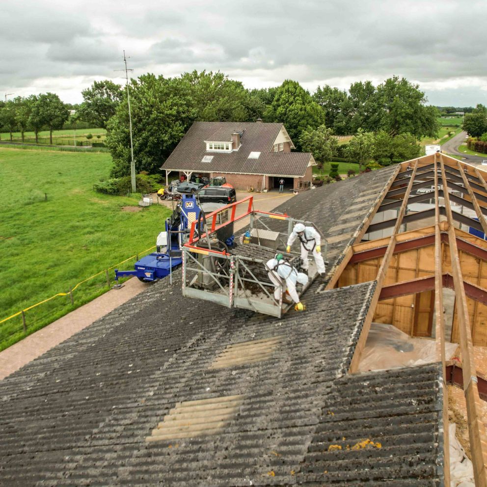 Twee arbeiders in beschermpakken op een hijsplatform verwijderen dakbedekking van groot landbouwschuurtje bij boerderij.