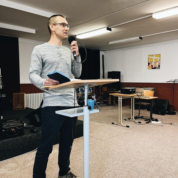 Man spreekt in microfoon achter lessenaar in een zaal, houdt notitieboekje, muziekinstrumenten en apparatuur op achtergrond.
