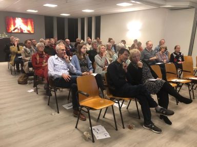 Groep volwassenen zit in rijen stoelen in een vergaderruimte, aandachtig luisterend naar een spreker.