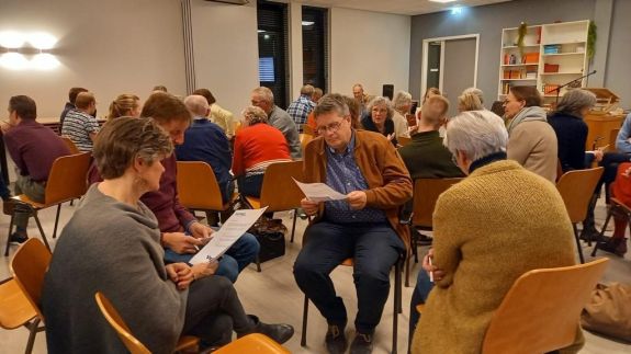 Groep mensen zit in kleine groepen op stoelen in een vergaderruimte, discussiërend en lezend uit papieren.