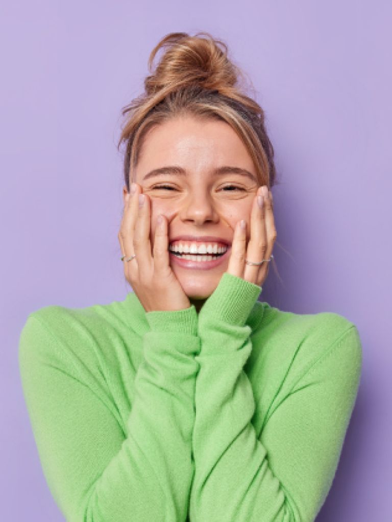 portrait-happy-young-female-model-keeps-hands-cheeks-smiles-broadly-shows-white-teeth-feels-glad-expresses-sincere-emotions-wears-green-turtleneck-isolated-purple-studio-background 1 Blonde mevrouw