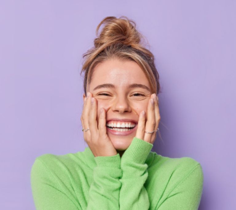 portrait-happy-young-female-model-keeps-hands-cheeks-smiles-broadly-shows-white-teeth-feels-glad-expresses-sincere-emotions-wears-green-turtleneck-isolated-purple-studio-background 1 Blonde mevrouw