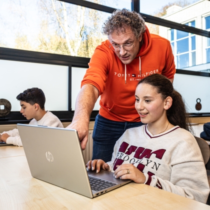 Foto van docent en leerlinge die samen naar een laptop scherm kijken. De docent wijst iets op het scherm aan en legt iets uit over waar ze samen naar kijken.