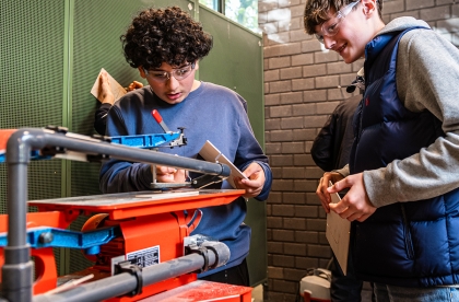 Twee leerlingen die een zaagapparaat bedienen. Ze hebben veiligheidsbrillen op.