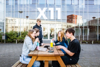Vier kinderen lunchen aan een picknicktafel voor de ingang van X11