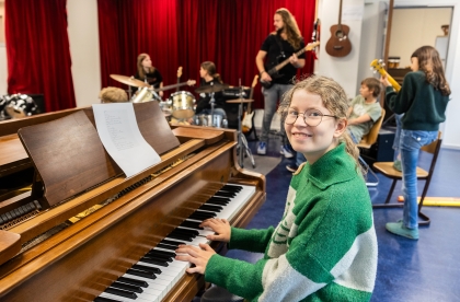 Decoratief. Leerling achter een piano. Op de achtergrond zijn meer leerlingen muziek aan het maken in een klaslokaal.