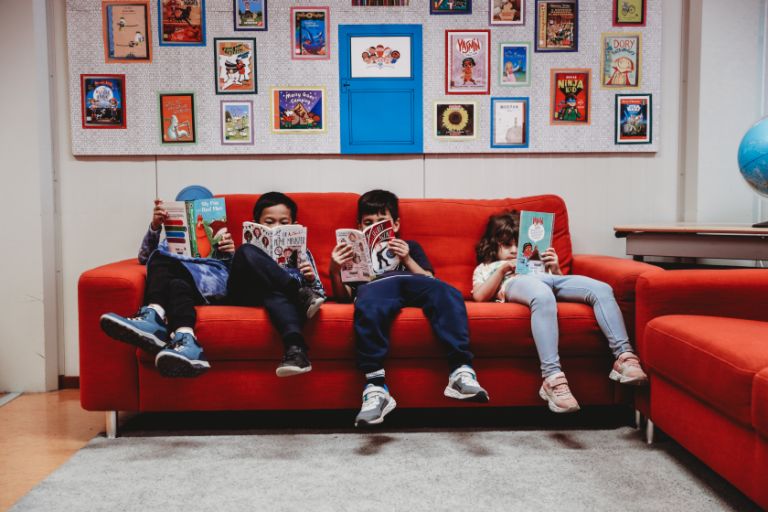 Four young pupils sit on a red couch at school, each reading a book Four young pupils sit on a red couch at school, each reading a book