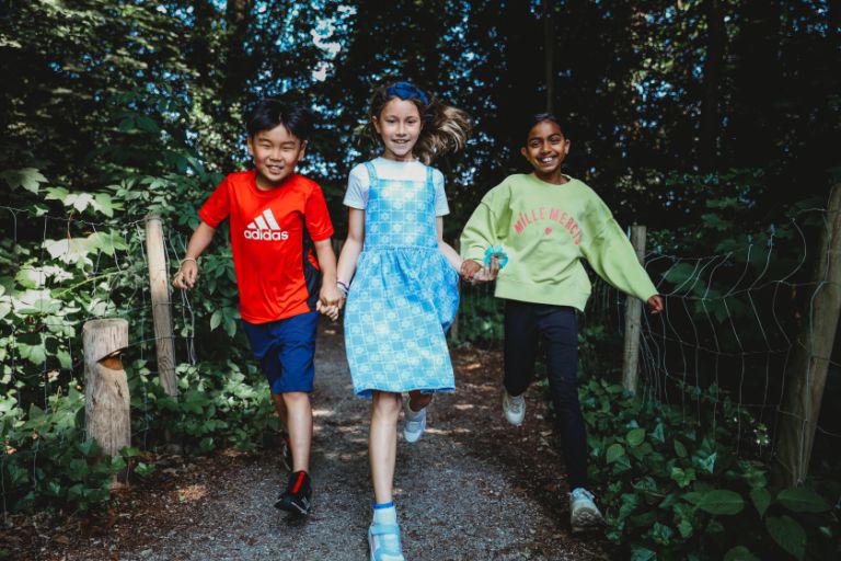 Three young pupils, smiling, holding hands and running down a pathway outdoors Three young pupils, smiling, holding hands and running down a pathway outdoors