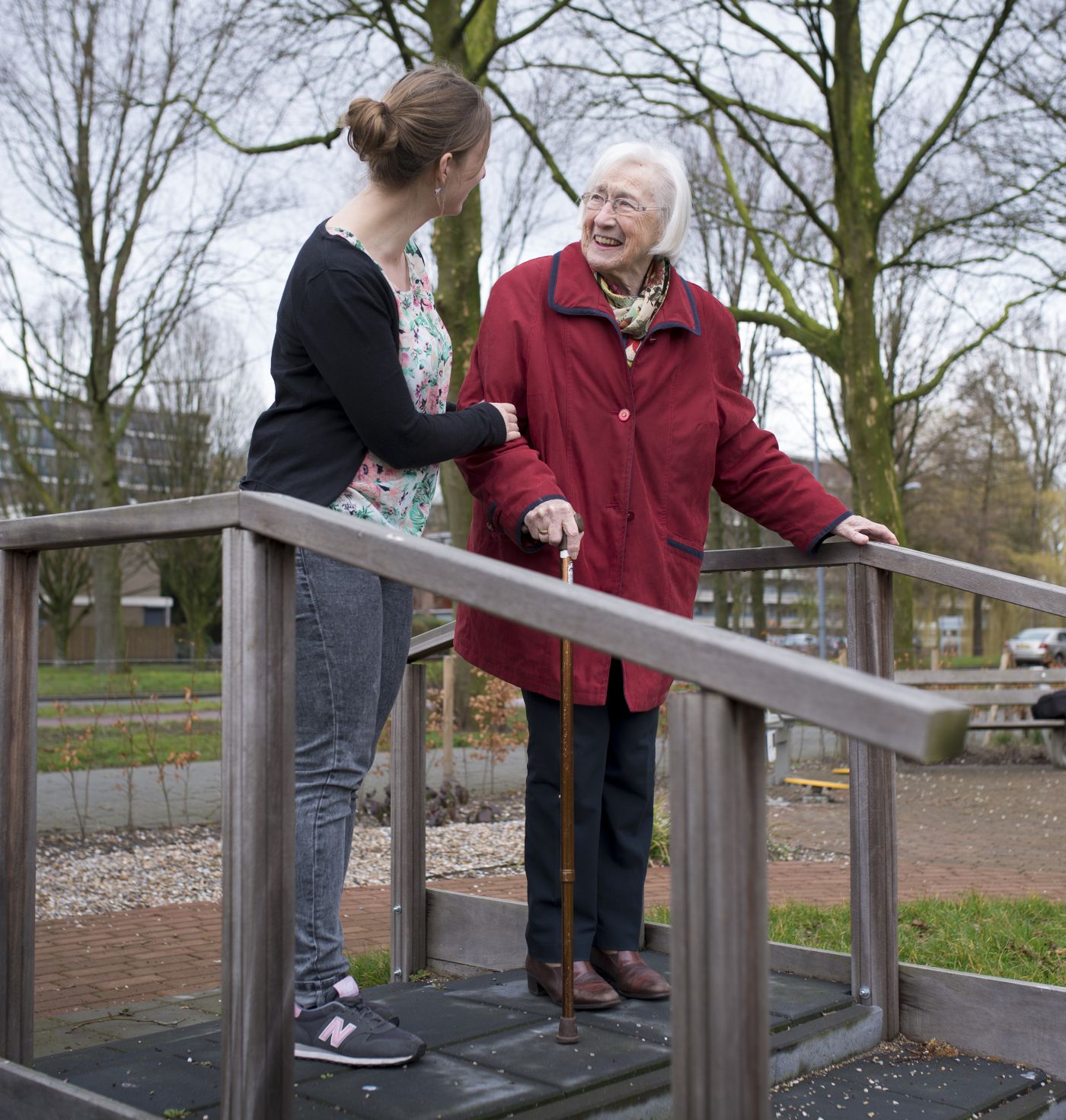 Fysiotherapeut en bewoonster lopen samen door beweegtuin bij verpleeghuis De Burcht in Rotterdam Het Lage Land Een oude vrouw en een jonge vrouw staan samen op een loopbruggetje in de tuin van een verpleeghuis en lachen naar elkaar