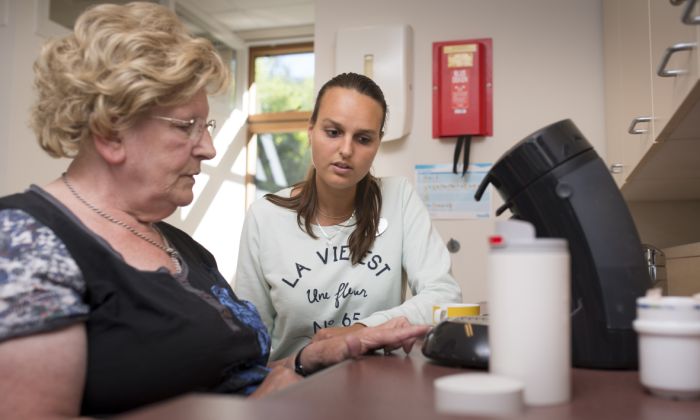 Ergotherapeut en cliënt van Lelie zorggroep in Rotterdam zetten een kopje koffie vacature behandeling ergotherapie Oudere vrouw en jongere vrouw kijken in een huiselijke omgeving naar een koffiezetapparaat en de oudere vrouw drukt op het knopje
