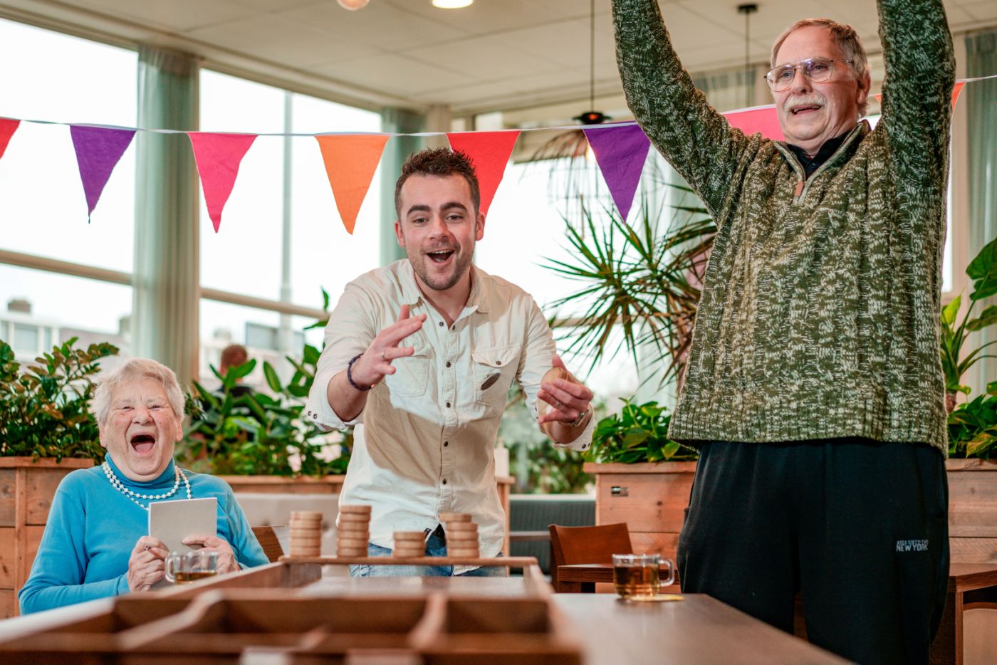 Activiteitenbegeleider sjoelt met cliënten in verpleeghuis Lelie zorggroep dagbesteding vacature vakantiewerk verpleeghuiszorg welzijn Mannelijke activiteitenbegeleider sjoelt met oude man en oude vrouw en sjoelen in verpleeghuis