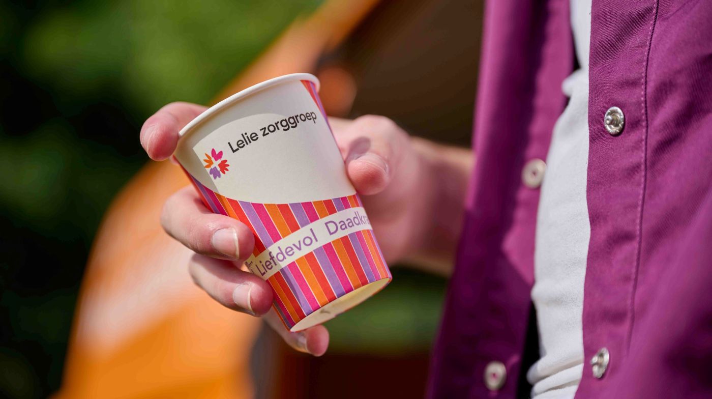 Beker drinken in hand met tekst liefdevol en daadkrachtig Man houdt gekleurd bekertje met de kerndaden van Lelie zorggroep vast waar de woorden Liefdevol en daadkrachtig zichtbaar zijn