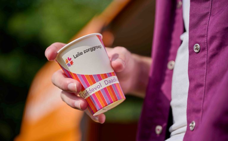 Beker drinken in hand met tekst liefdevol en daadkrachtig Man houdt gekleurd bekertje met de kerndaden van Lelie zorggroep vast waar de woorden Liefdevol en daadkrachtig zichtbaar zijn