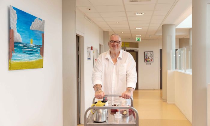 Bewoner van Korsakov Expertisecentrum Slingedael loopt de koffieronde Meneer in witte blouse staat in een gang achter karretje met koffiekannen en een koektrommel