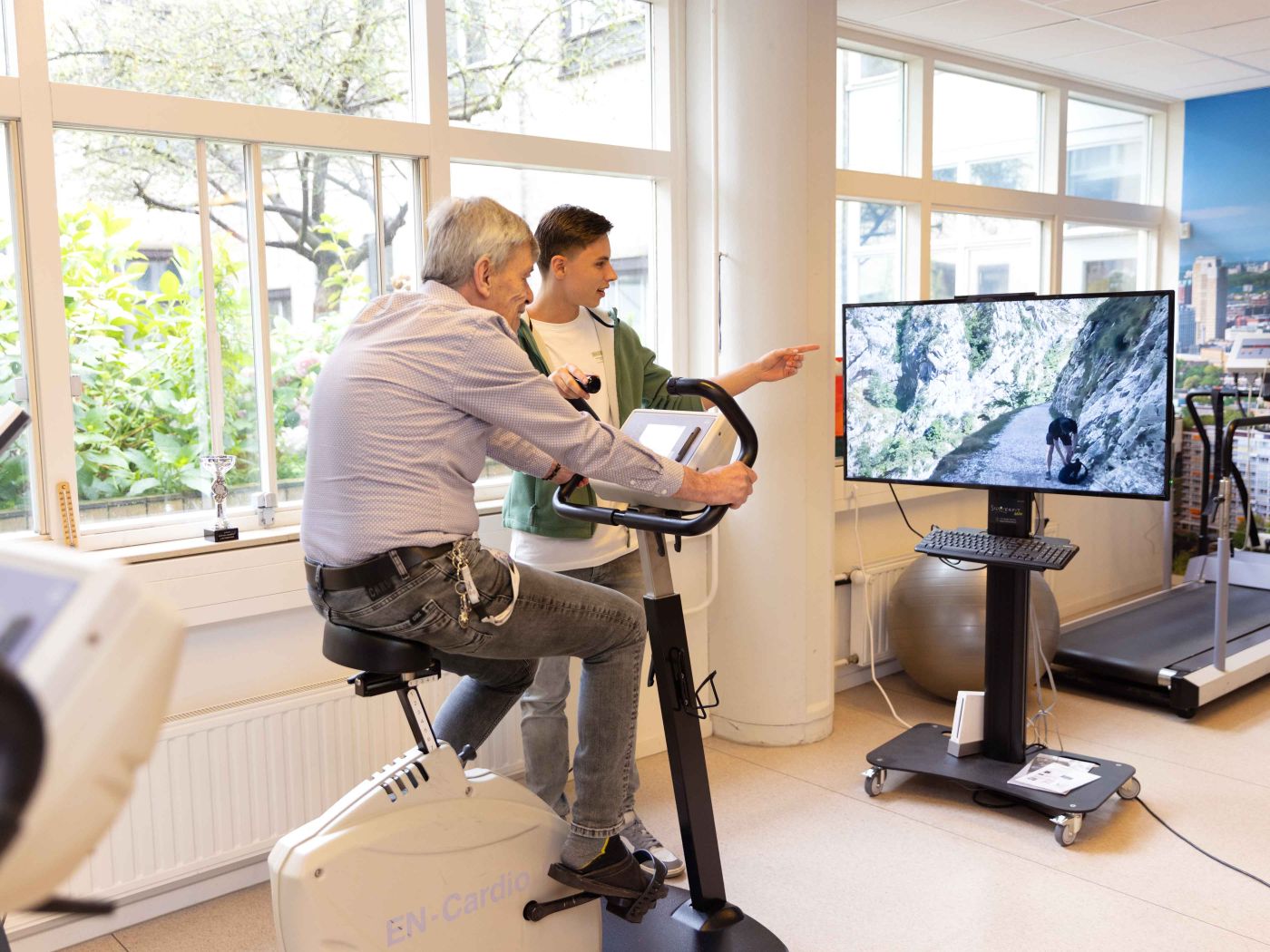 Bewoner krijgt fysiotherapie in de oefenruimte van Korsakov Expertisecentrum Slingedael in Rotterdam Meneer zit op een hometrainer in de fysioruimte van Slingedael en kijkt naar een scherm waarop een mooi landschap te zien is, terwijl een medewerker van Slingedael hem aanwijzingen geeft