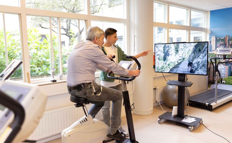 Bewoner krijgt fysiotherapie in de oefenruimte van Korsakov Expertisecentrum Slingedael in Rotterdam vacature fysiotherapeut behandeling verpleeghuiszorg Meneer zit op een hometrainer in de fysioruimte van Slingedael en kijkt naar een scherm waarop een mooi landschap te zien is, terwijl een medewerker van Slingedael hem aanwijzingen geeft