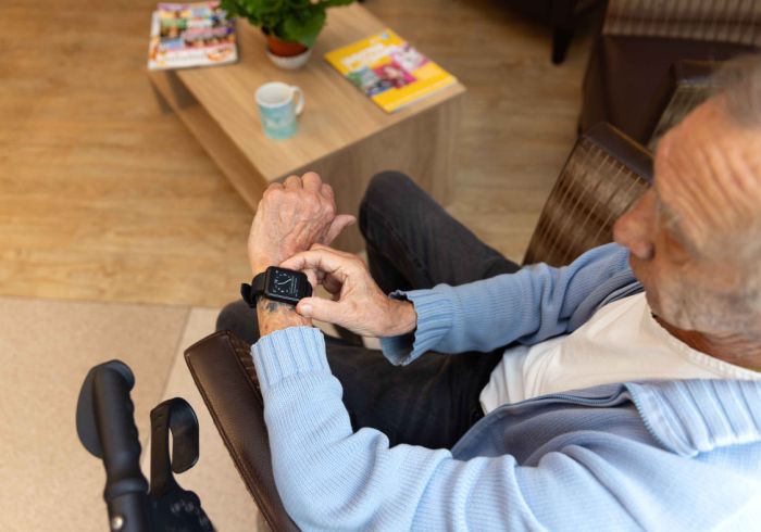 Meneer bekijkt zijn MyWebb beeldhorloge in Korsakov Expertisecentrum Slingedael in Rotterdam Meneer in een blauw vest zit in een fauteuil en kijkt op een elektronisch horloge