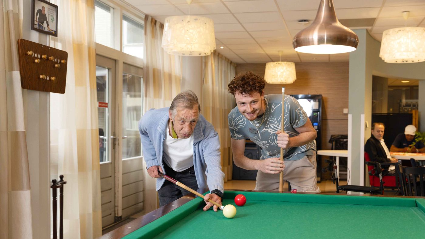 Bewoner en medewerker van Korsakov Expertisecentrum Slingedael spelen een potje biljart bij de biljarttafel in de ontmoetingsruimte vacature welzijn dagbesteding vrijwilliger Oudere man en jonge man spelen samen een potje biljart en buigen zich over de tafel om de keu goed te kunnen richten