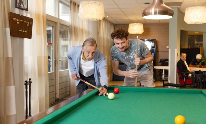 Bewoner en medewerker van Korsakov Expertisecentrum Slingedael spelen een potje biljart bij de biljarttafel in de ontmoetingsruimte vacature welzijn dagbesteding vrijwilliger Oudere man en jonge man spelen samen een potje biljart en buigen zich over de tafel om de keu goed te kunnen richten