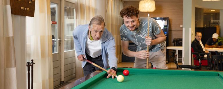 Bewoner en medewerker van Korsakov Expertisecentrum Slingedael spelen een potje biljart bij de biljarttafel in de ontmoetingsruimte vacature welzijn dagbesteding vrijwilliger Oudere man en jonge man spelen samen een potje biljart en buigen zich over de tafel om de keu goed te kunnen richten