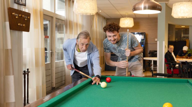 Bewoner en medewerker van Korsakov Expertisecentrum Slingedael spelen een potje biljart bij de biljarttafel in de ontmoetingsruimte vacature welzijn dagbesteding vrijwilliger Oudere man en jonge man spelen samen een potje biljart en buigen zich over de tafel om de keu goed te kunnen richten