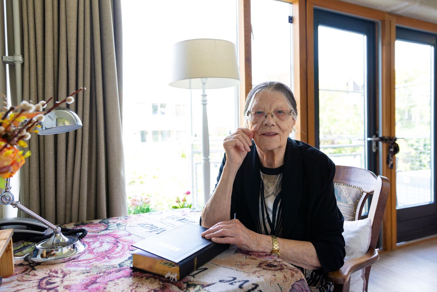 Bewoner aan tafel met de Bijbel en kijkt in camera Oude mevrouw zit aan tafel in haar slaapkamer met een bijbel op tafel