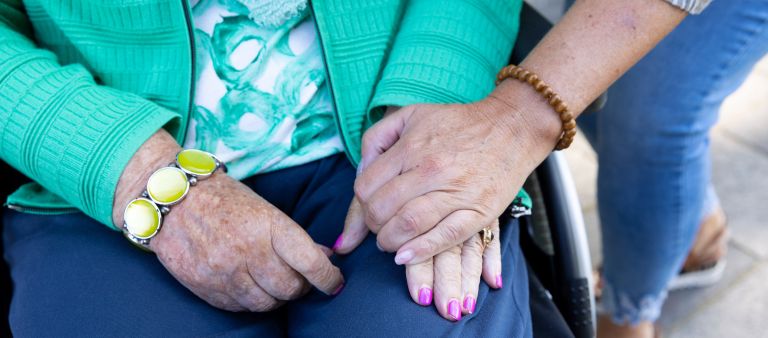 Bewoner en mantelzorger houden elkaars hand vast bij verpleeghuis Tiendhove Oude en jongen handen op elkaar op schoot