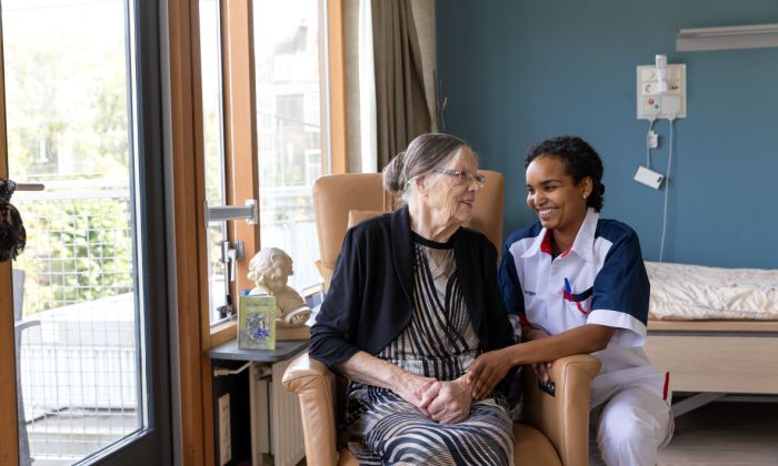 Bewoner en zorgverlener in stoel op eigen kamer vacature zorg Pniël verpleeghuiszorg Oude mevrouw zit in stoel en zorgmedewerker zit gehurkt naast mevrouw in haar appartement in het verpleeghuis