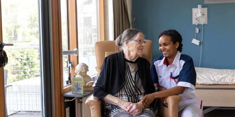 Bewoner en zorgverlener in stoel op eigen kamer vacature zorg Pniël verpleeghuiszorg Oude mevrouw zit in stoel en zorgmedewerker zit gehurkt naast mevrouw in haar appartement in het verpleeghuis