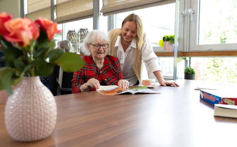 Bewoner kleinkind mantelzorger lezen verpleeghuis vacature welzijn Pniël dagbesteding vrijwilliger Oude mevrouw zit aan tafel en leest een tijdschrift en kleinkind leest over de schouder mee