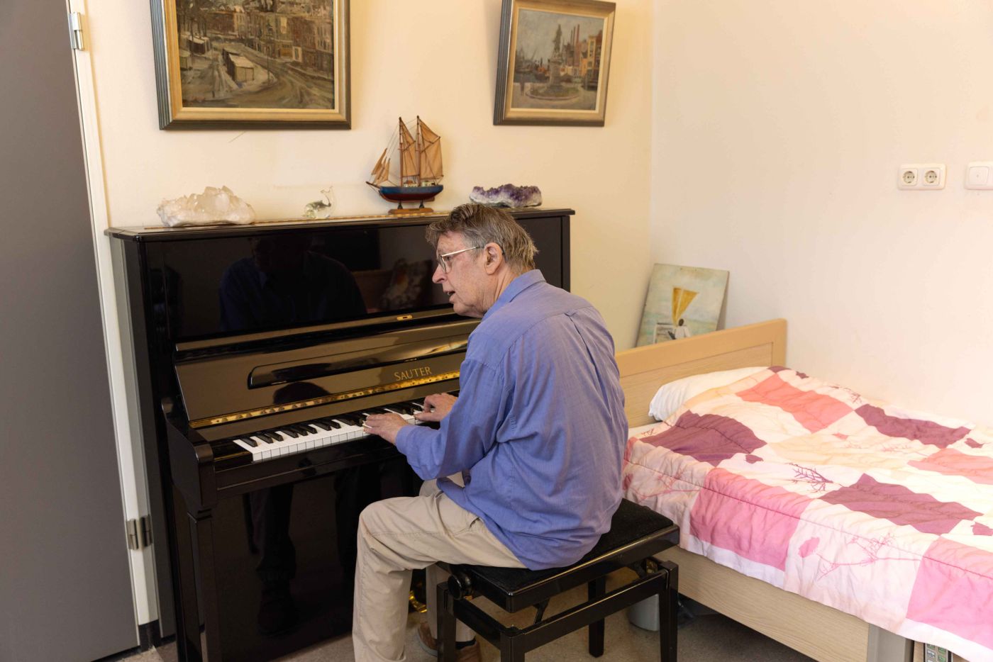 Bewoner van Korsakov Expertisecentrum Slingedael achter de piano in zijn eigen kamer Meneer in blauwe blouse speelt op een piano in zijn eigen kamer bij Slingedael
