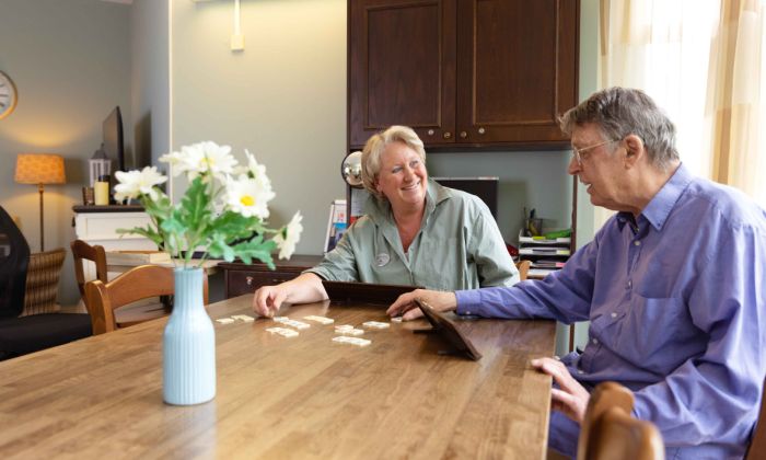 Zorgmedewerker en bewoner doen een spelletje Rummikub in Korsakov Expertisecentrum Slingedael vacature welzijn Man en vrouw zitten samen aan een tafel in een huiskamer en spelen lachend een spelletje Rummikub