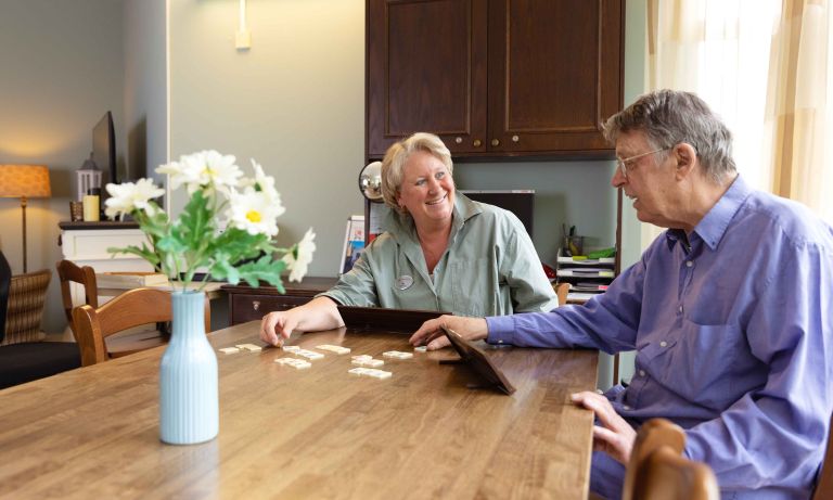 Zorgmedewerker en bewoner doen een spelletje Rummikub in Korsakov Expertisecentrum Slingedael vacature welzijn Man en vrouw zitten samen aan een tafel in een huiskamer en spelen lachend een spelletje Rummikub