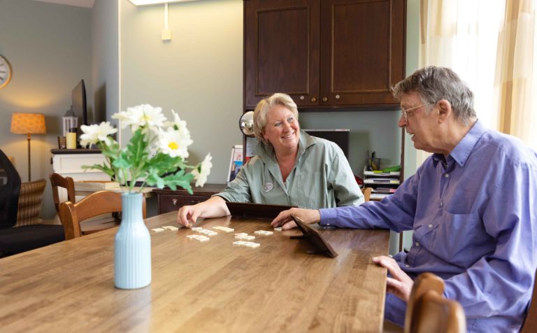 Zorgmedewerker en bewoner doen een spelletje Rummikub in Korsakov Expertisecentrum Slingedael vacature welzijn Man en vrouw zitten samen aan een tafel in een huiskamer en spelen lachend een spelletje Rummikub