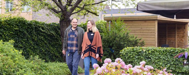 Bewoner en medewerker van Korsakov Expertisecentrum Slingedael lopen een rondje in de tuin vacature sph social work hulpverlening verpleeghuiszorg Oudere man en jongere vrouw lopen naast elkaar in de tuin van Slingedael en lachen naar elkaar