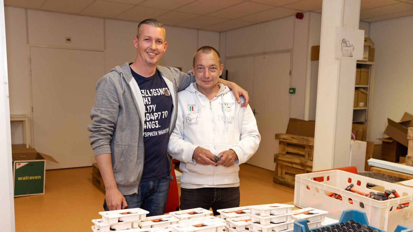 Bewoner werkt samen met medewerker bij de werkplaats van Korsakov Expertisecentrum Slingedael in Rotterdam welzijn vacature dagbesteding Twee mannen staan bij een werktafel in de werkplaats van Slingedael en de ene man heeft zijn arm om de schouder van de andere man geslagen