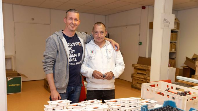 Bewoner werkt samen met medewerker bij de werkplaats van Korsakov Expertisecentrum Slingedael in Rotterdam welzijn vacature dagbesteding Twee mannen staan bij een werktafel in de werkplaats van Slingedael en de ene man heeft zijn arm om de schouder van de andere man geslagen