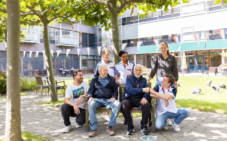 Bewoners en zorgverleners op en rondom een bankje in de tuin Vier zorgmedewerkers staan bij een bankje met een mannelijke client en familieleden.