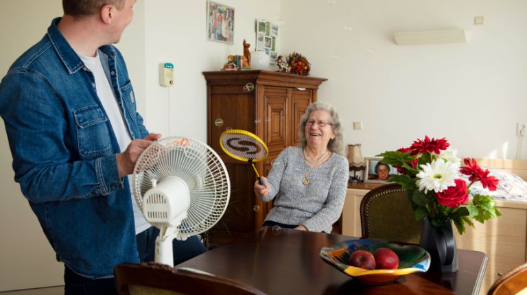Bewegingsagoog en bewoner spelen spelletje in christelijk verpleeghuis De Burcht in Rotterdam Het Lage Land Jonge man en oude vrouw staan rond een ventilator en de vrouw slaat met een racket zeepbellen weg die de ventilator wegblaast.