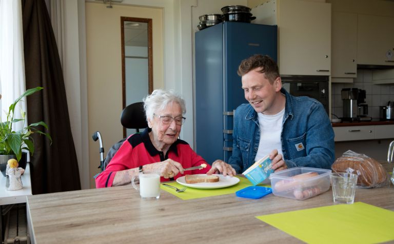 Bewegingsagoog en bewoner smeren samen een boterham in verpleeghuis De Burcht in Rotterdam vacature welzijn bewegingsagogiek behandeling ergotherapie dietetiek fysiotherapie Jonge man en oudere vrouw zitten samen aan tafel en de vrouw smeert zelf een boterham met een mes terwijl de jongen lachend toekijkt
