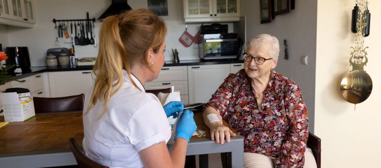 Cliënt arm op tafel zorgverlener verbinden in keuken vacature thuiszorg uniform Cliënt arm op tafel zorgverlener verbinden in keuken vacature