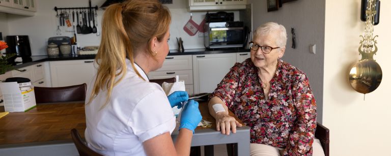 Cliënt arm op tafel zorgverlener verbinden in keuken Vrouwelijke zorgverlener verbindt de arm van een oude vrouw op tafel in de keuken van het huis
