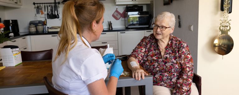 Cliënt arm op tafel zorgverlener verbinden in keuken Vrouwelijke zorgverlener verbindt de arm van een oude vrouw op tafel in de keuken van het huis