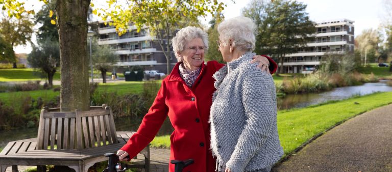 Cliënt en mantelzorger arm om schouder buiten Een oude dame loopt met een rollater samen met andere oude mevrouw die een arm om haar schouder slaat