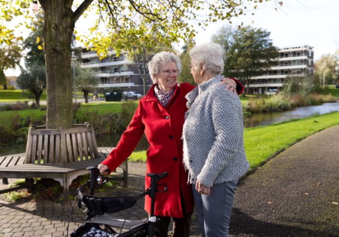 Cliënt en mantelzorger arm om schouder buiten Oude vrouw achter rollator staat buiten in een groene omgeving met mantelzorger die een arm om schouder legt