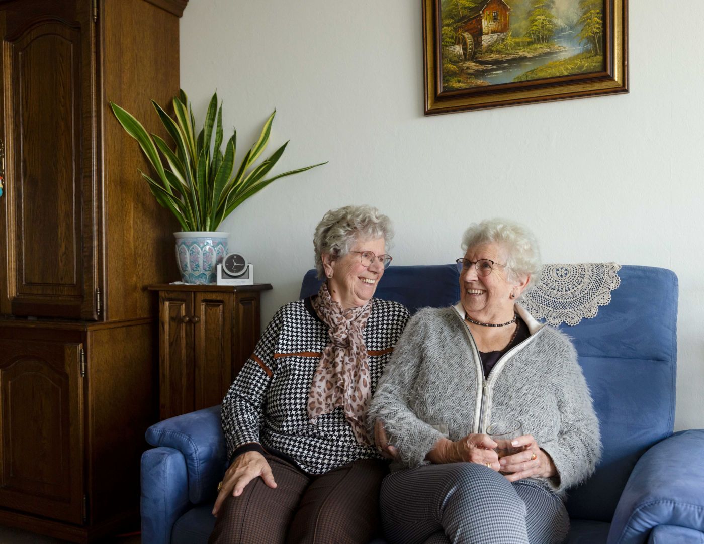 Cliënt en mantelzorger lachend bank huiskamer Vrouwelijke cliënt en zus als mantelzorger zitten samen in de huiskamer naast elkaar op de bank te praten en te lachen