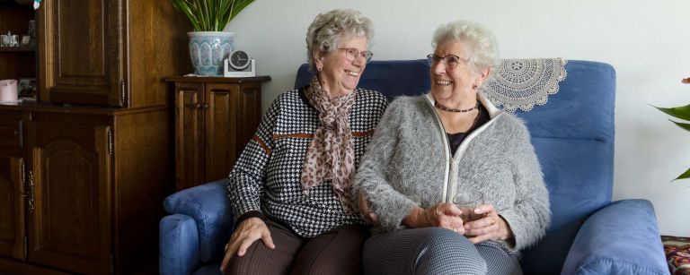 Cliënt en mantelzorger lachend bank huiskamer Vrouwelijke cliënt en zus als mantelzorger zitten samen in de huiskamer naast elkaar op de bank te praten en te lachen