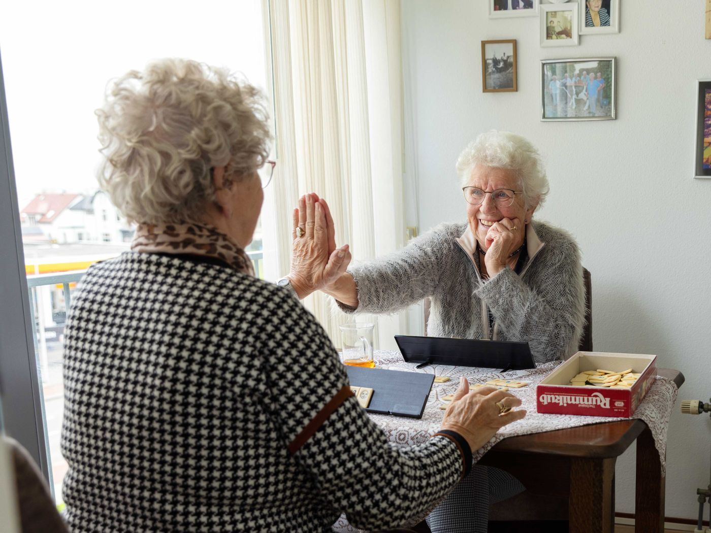 Cliënt en mantelzorger rummikub high five Oudere dames spelen een spelletje Rummikub aan tafel in de woonkamer en geven elkaar een high five