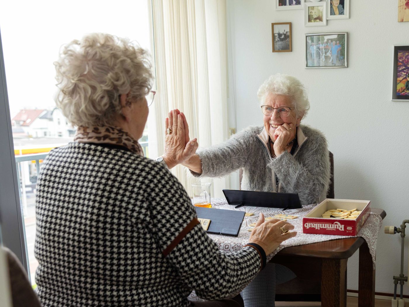 Cliënt en mantelzorger rummikub high five Oudere dames spelen een spelletje Rummikub aan tafel in de woonkamer en geven elkaar een high five